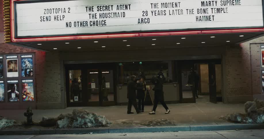 The video shows people standing and walking in front of a movie theater entrance during a cold evening a large marquee above the doors lists several upcoming film titles including zootopia 2 and marty supreme