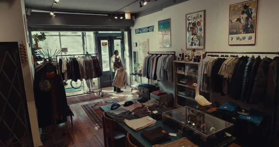 A person browses through a rack of clothing near the entrance of a boutique shop the interior is filled with various garments framed posters on the walls and a decorative wooden goose on a display table