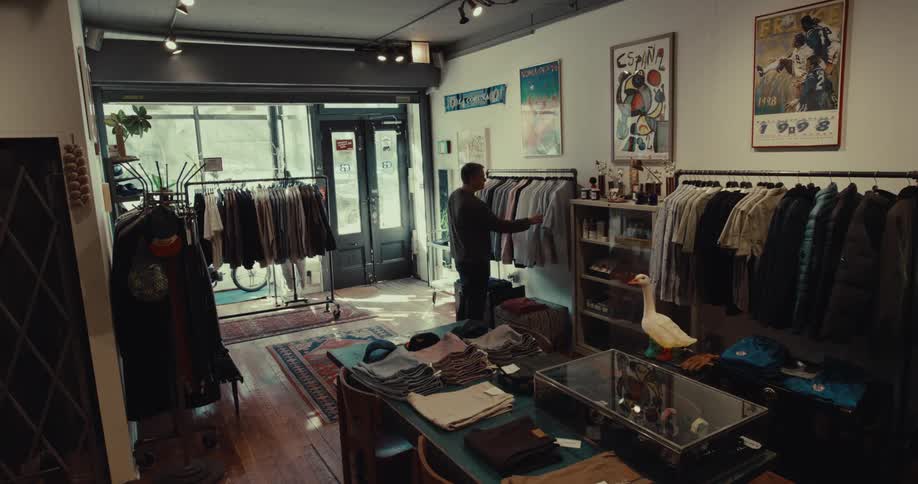 A man walks into a vintage clothing shop and begins browsing through various garments hanging on metal racks near a wooden table and a glass display case containing watches