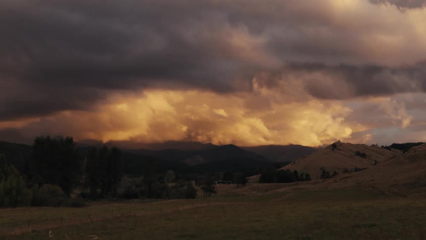 Dark storm clouds hang over a vast mountain range while golden sunset light glows through a central opening in the sky above a quiet valley with rolling hills and scattered trees