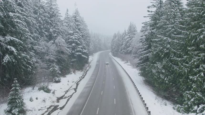 A white SUV travels along a snowy mountain pass highway surrounded by a forest of evergreen trees