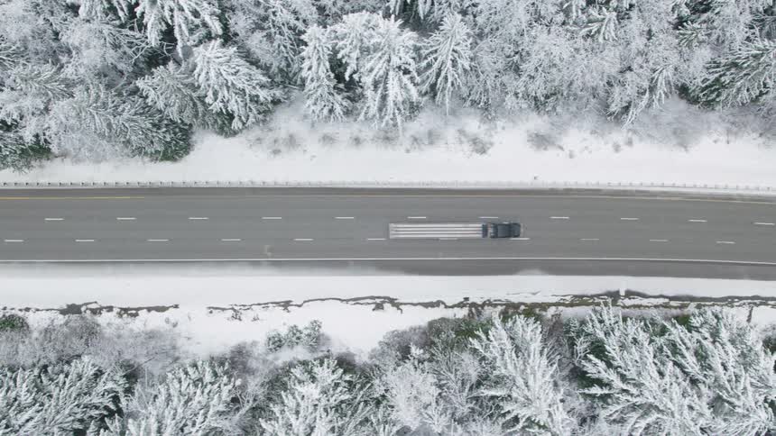 Parallel traverse above a semi truck traveling on a highway between snowcovered trees amidst light flurries