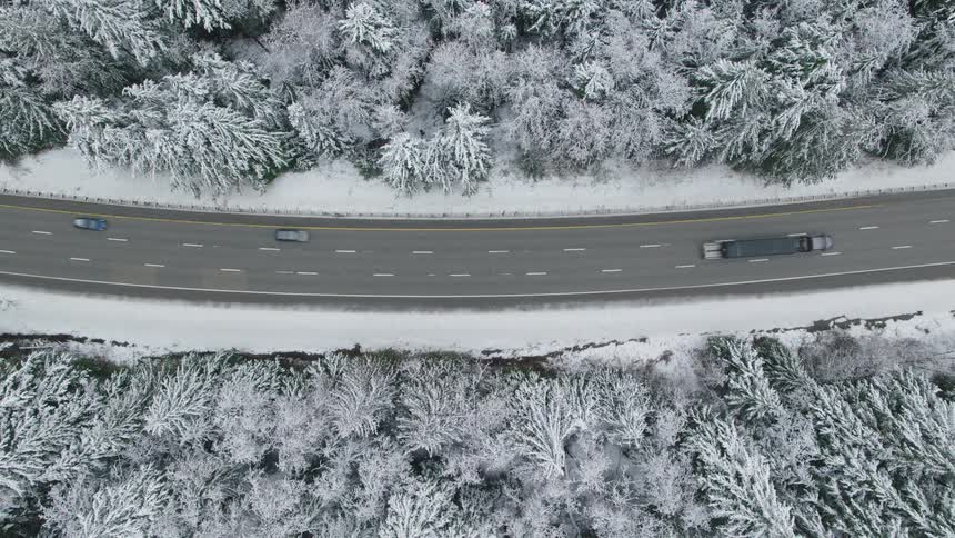 Cars and semi trucks moves along a curved section of freeway through a snow covered forest