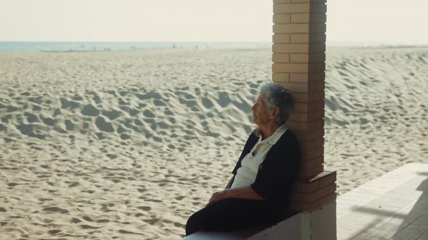 Elderly woman sitting against a column looking toward a sandy beach in daylight
