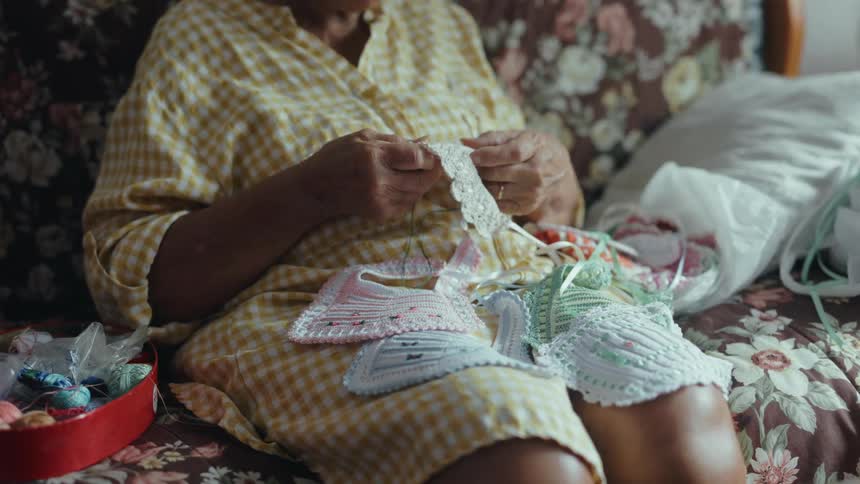 An elderly person sits on a floral sofa while carefully sewing or mending small white lace garments