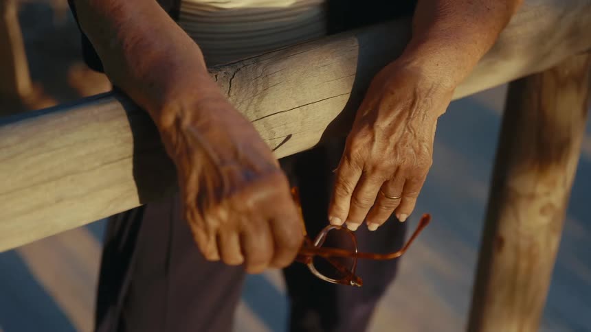The video features a closeup of an elderly persons weathered hands resting on a wooden railing during golden hour one hand holds a pair of glasses while the other rests still highlighting the detailed texture of the skin and a simple gold