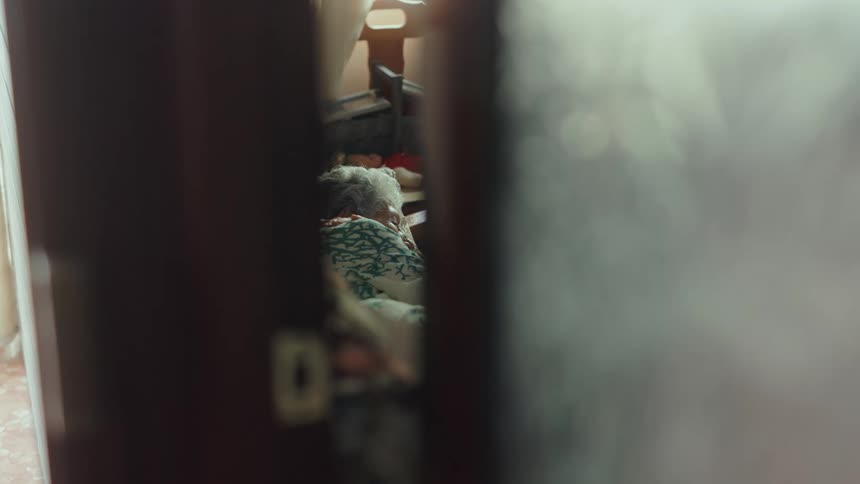 Elderly woman sleeps in bed seen through a door gap