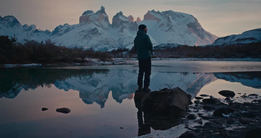 Person standing on a rock by a lake reflecting snowcovered mountains