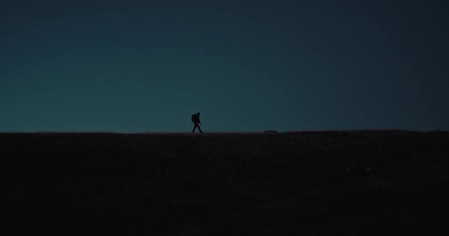 A lone figure wearing a backpack and headlamp walks across a dark ridge against a deep blue twilight sky the silhouette moves steadily along the horizon before stopping to look directly toward the camera