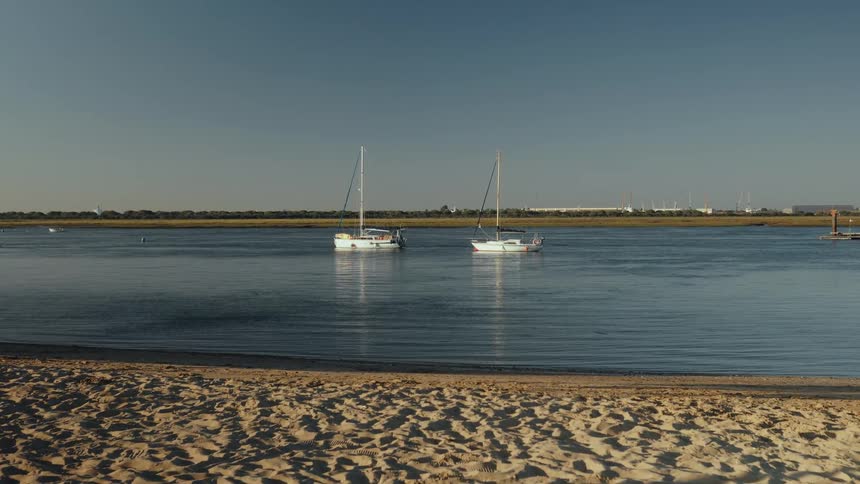 Two sailboats anchored on calm water near a sandy beach with a distant shoreline