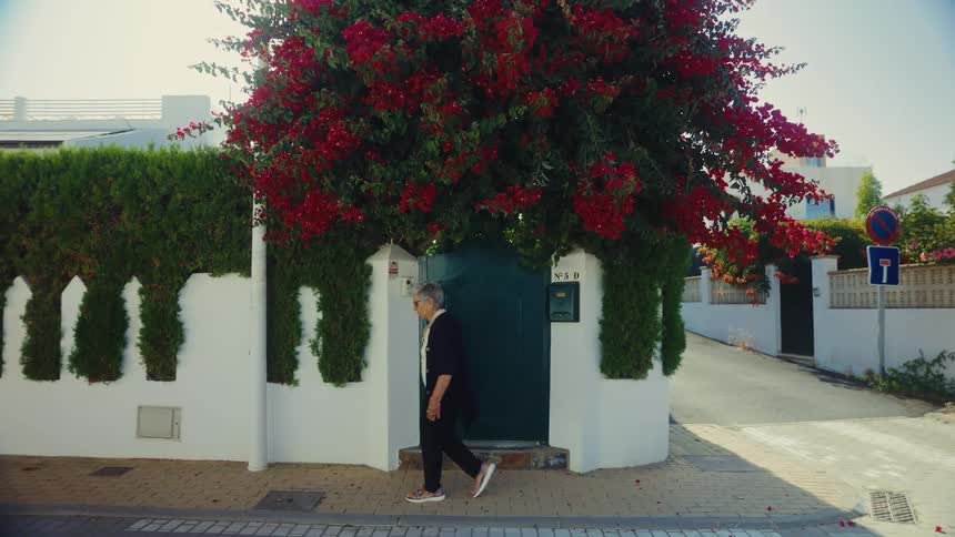 Elderly person walking in front of a green gate under a tree with red flowers