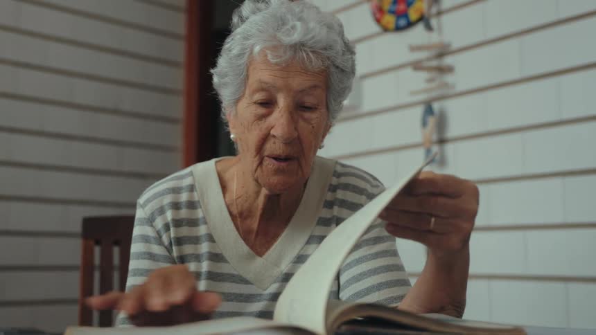 An elderly woman with gray hair sitting behind a desk flipping the page of a book and wearing a wedding ring