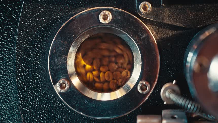Coffee beans visible through a circular metal viewing window in a roasting machine