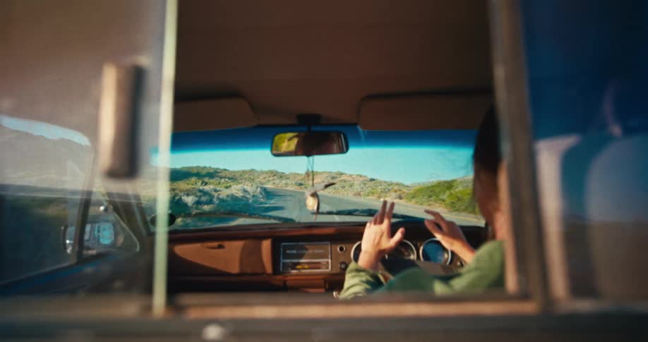 A person drives a vintage vehicle along a coastal road toward sunlit hills during golden hour