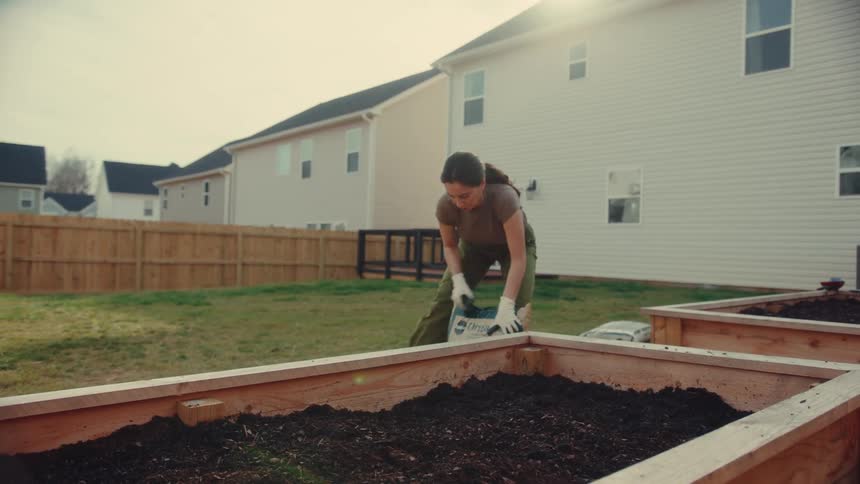 Woman pouring a bag of soil into a wooden raised garden bed in a backyard