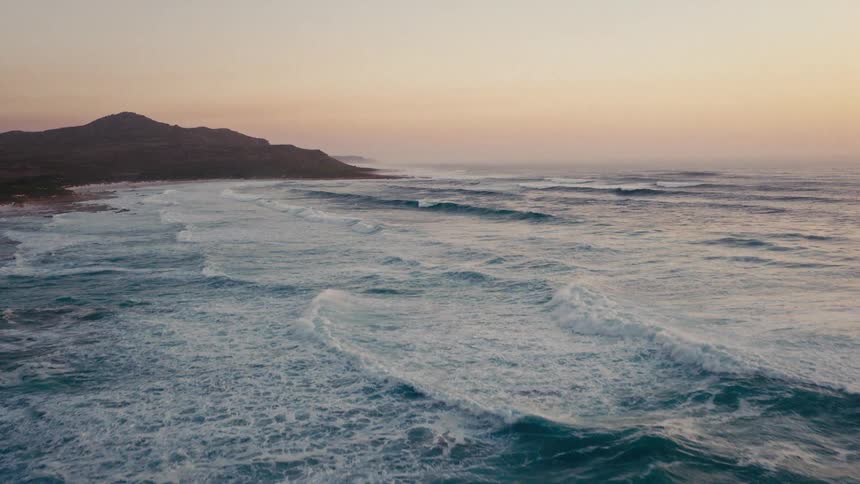 Ocean waves moving toward the coast with mountains in the background at sunset