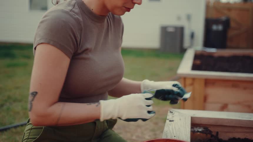 A woman wearing gardening gloves sits beside a wooden raised bed and carefully examines a packet of seeds she then uses a hand trowel to prepare the dark fresh soil for planting while other seed packets wait in a nearby wooden bowl