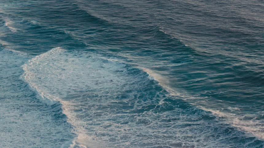 Ocean waves swell and break into white foam across the deep blue surface of the water during a wide high angle shot
