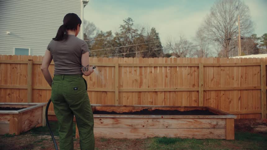 A woman uses a handheld water spray nozzle to water soil in a raised garden bed in a backyard