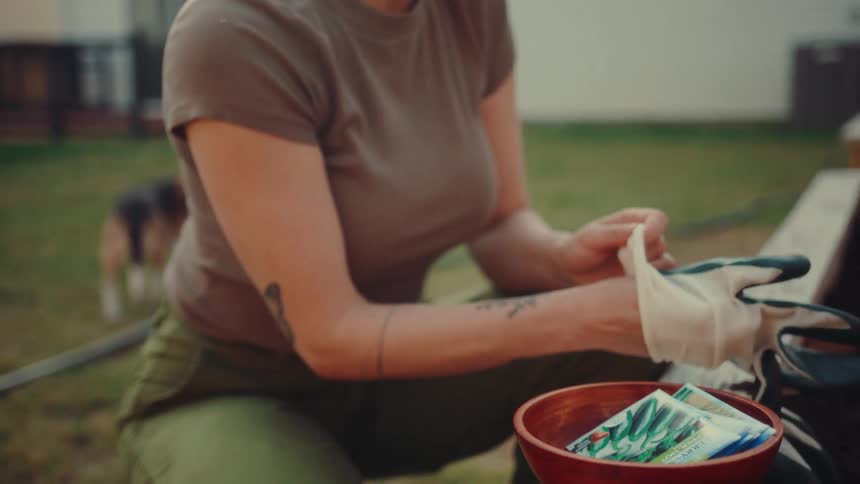 A woman sits beside a raised garden bed in her backyard looking through several colorful seed packets before placing them in a wooden bowl and pulling on a pair of white and black gardening gloves to begin planting in the dark soil