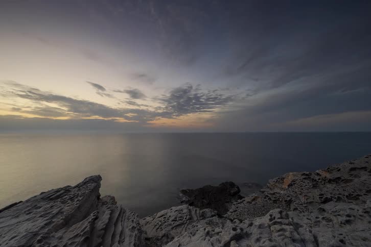 Jagged grey cliffs overlook a calm ocean under a darkening sky where orange sunset light glows through scattered clouds