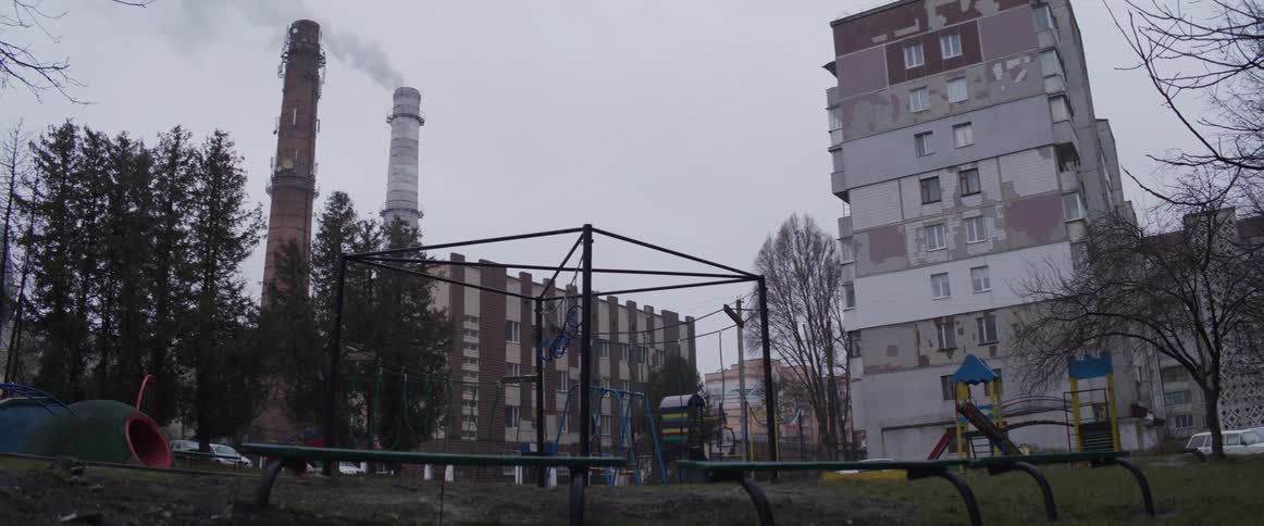 An apartment building stands next to a playground with metal swings and a slide while two tall industrial chimneys emit smoke into a gray overcast sky in a quiet urban neighborhood