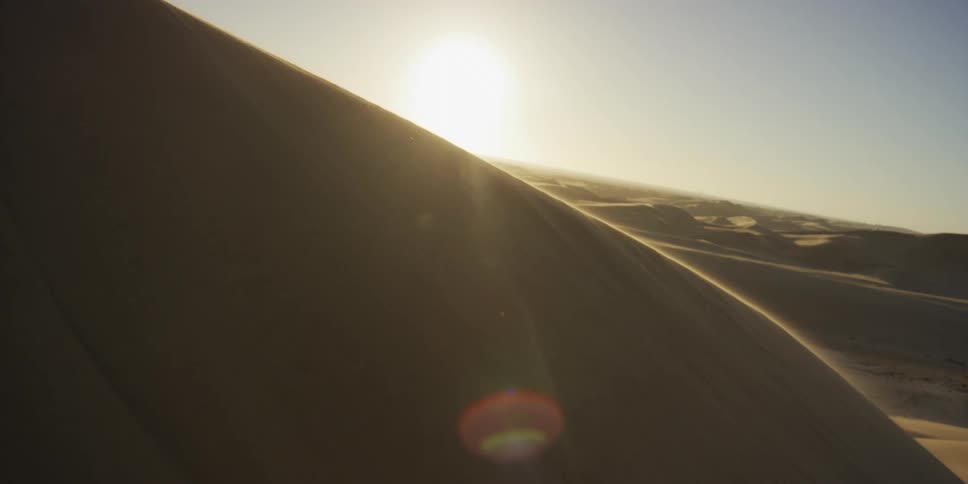 The sun hangs low over a vast desert landscape where wind blows fine sand across the crest of a steep dune creating a golden haze against the clear sky and casting long shadows over the undulating ground