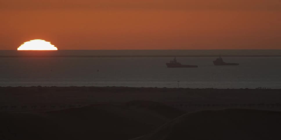 Boats float in the water off the beach as the sun sets in the red sky