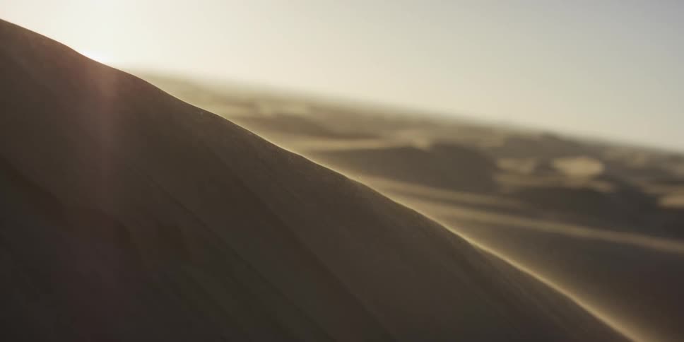 Fine sand blows off the crest of a steep desert dune as warm sunlight glows across the vast arid landscape during the late afternoon