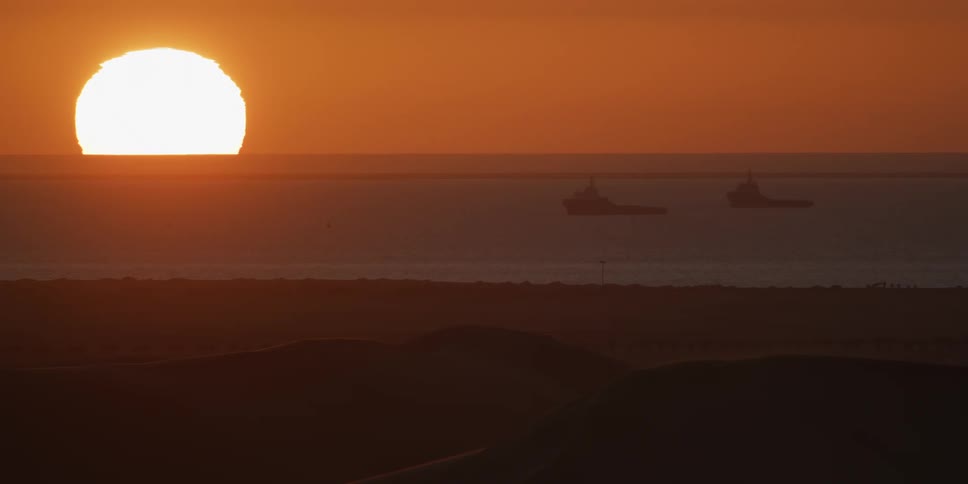 Sun rises on the horizon with ships at sea behind dunes