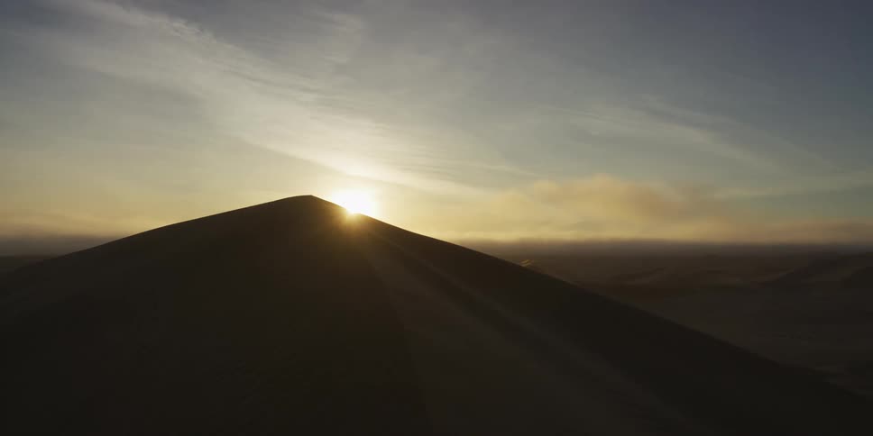 A cinematic drone shot captures the sun rising behind a massive sand dune in a vast desert