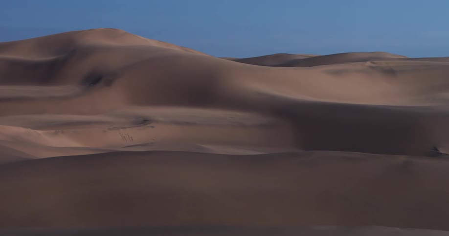 Large brown sand dunes roll across a vast desert landscape under a clear blue sky while soft shadows define the curved ridges and distant peaks of the arid wilderness