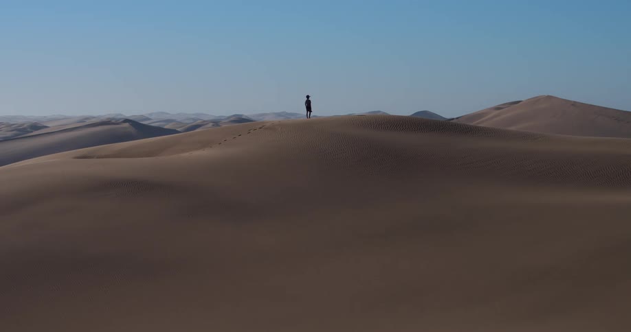 A person runs up a large sand dune that overlooks a massive desert
