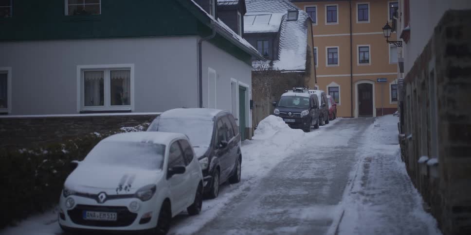 A white hatchback and several other cars sit parked along a narrow cobblestone street covered in slush and snow between colorful residential buildings