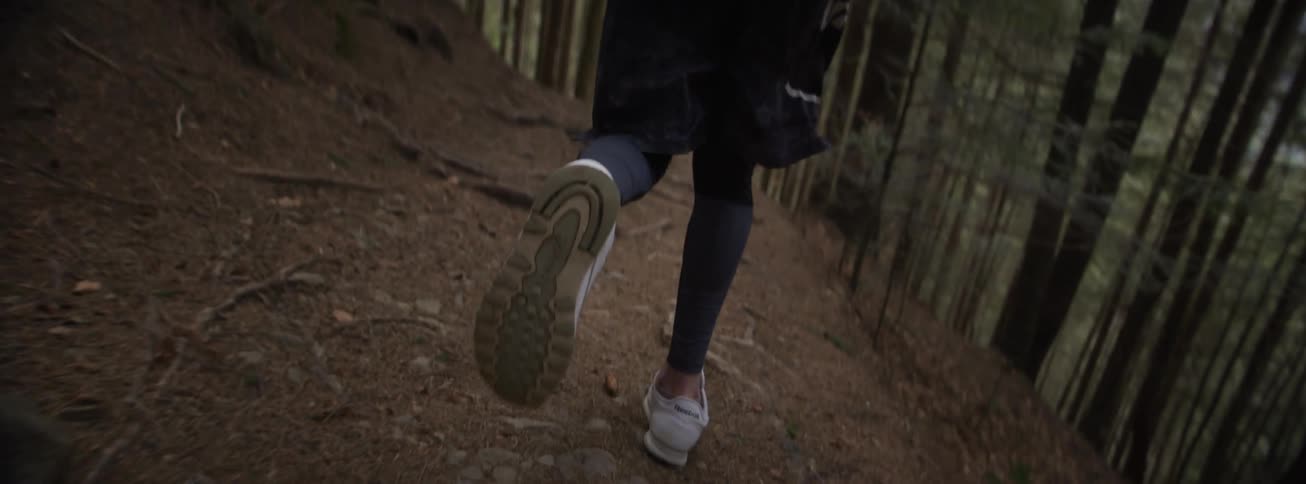A close view from behind shows the legs of a man wearing leggings and white sneakers as he runs up a dirt path through a dense pine forest with tall trees in the background