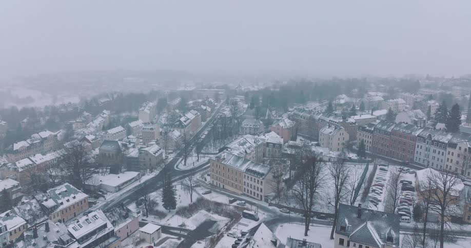 A town covered in white snow under a heavy gray sky showing rows of multi story houses winding roads with a few vehicles and clusters of bare trees scattered