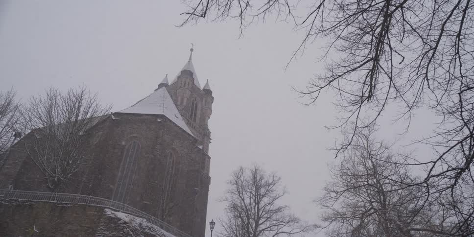 A stone church tower rises into a heavy snowfall partially framed by the bare branches of winter treesthe scene captures a steady descent of snowflakes against the backdrop of the buildings gothic architecture and a gray overcast sky
