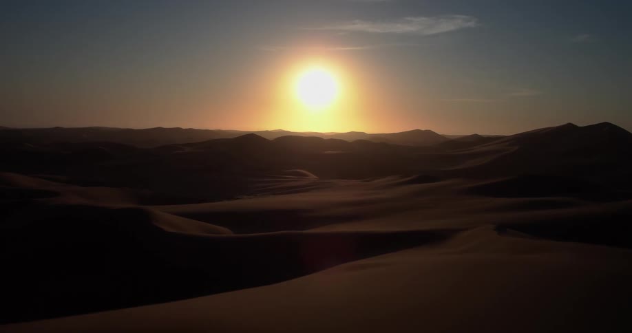 A group of people stands silhouetted on the crest of a dark sand dune as a bright sun sets over a vast desert landscape filled with rolling hills and distant mountains under a clear sky