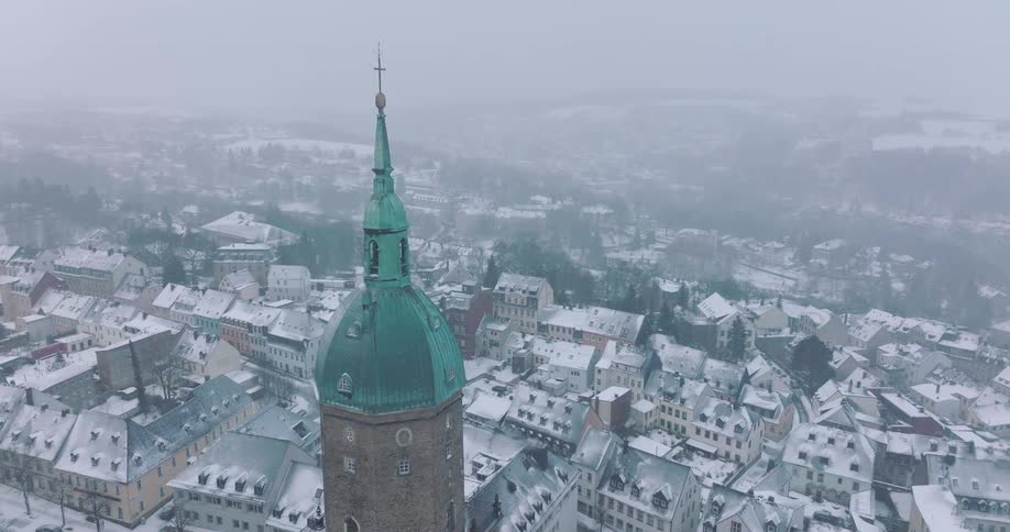 An aerial view shows the green dome and copper spire of saint annen church tower overlooking the snow covered rooftops of annaberg buchholz during a foggy winter day