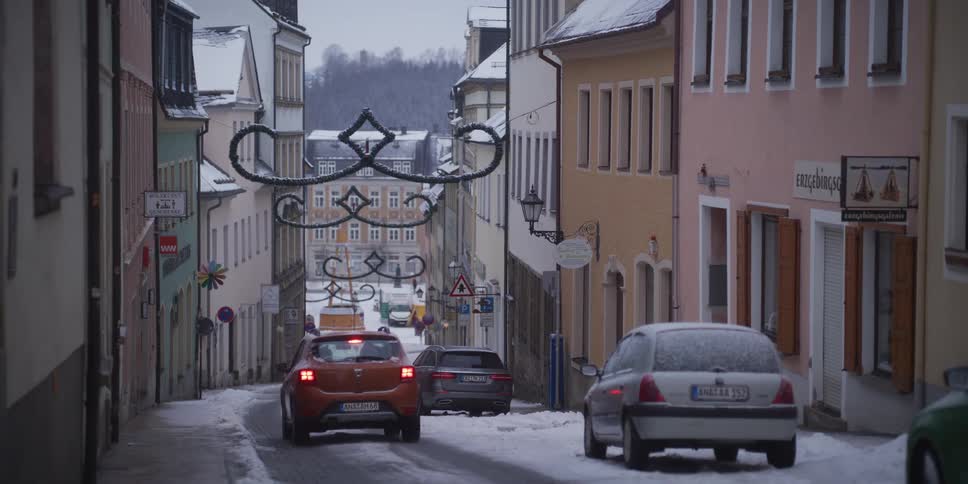 An orange car drives down a narrow snow covered street lined with pastel colored buildings and festive holiday decorations hanging overhead on a cold winter day