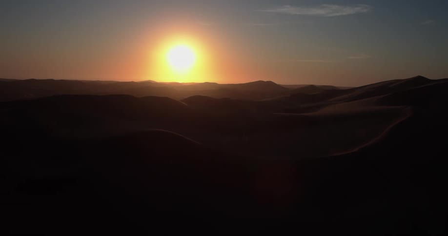 A wide view captures the bright golden sun hanging low over the rolling dunes of the sahara desert as the sunlight creates deep shadows across the vast and undulating sand during a warm sunset
