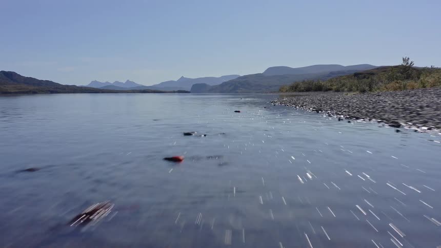 A drone flies low over a rocky shoreline and clear blue water in a river valley with distant mountains and a vast treeless landscape under a bright sunny sky