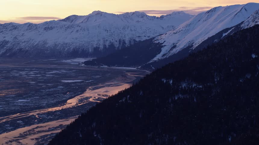 The video displays a vast snowcovered mountain range overlooking a winding river valley during a cold winter sunsetthe fading light casts deep purple shadows across the forested slopes while reflecting a warm golden glow off the icy wat