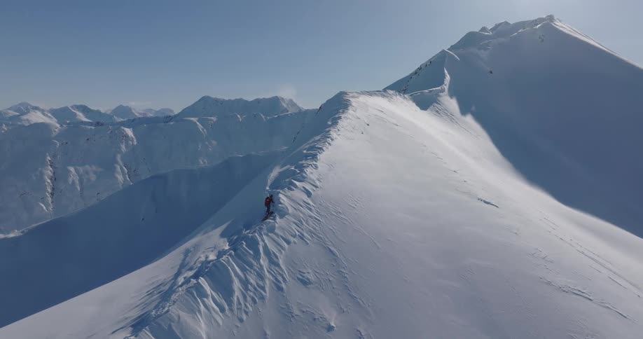 A man wearing a red jacket and a backpack walks carefully along a narrow snow ridge on a high mountain peak under a clear blue sky during a bright winter day