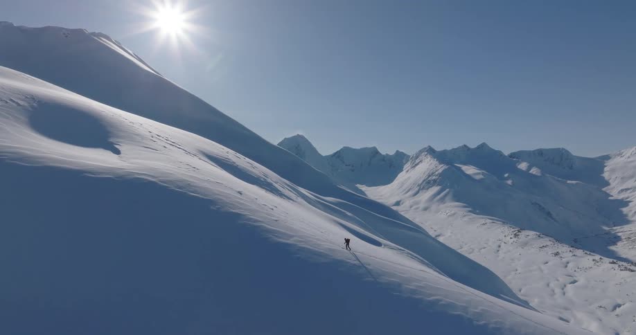 Mountaineer holds his trekking poles as he climbs up a snow covered mountain slope with mountains in the distance