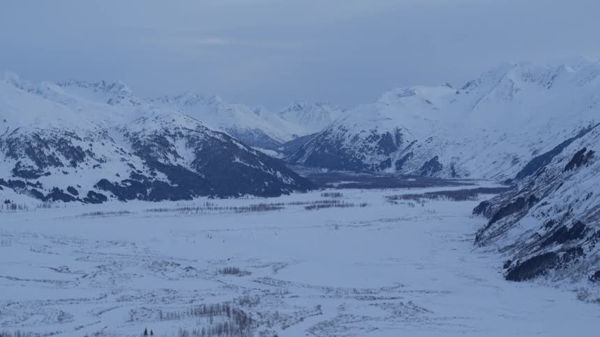 A wide view shows the snow covered peaks of the chugach mountains surrounding a vast frozen valley with sparse trees under a blue twilight sky in alaska