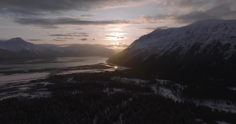 An aerial view shows the sun setting behind the snow covered chugach mountains in alaska above a vast valley and a dark evergreen forest during winter