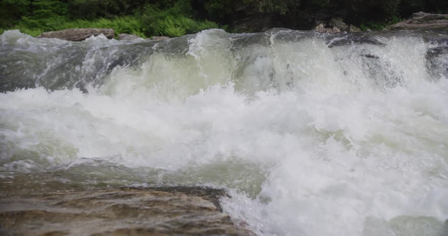 A close view captures the powerful rush of water as it tumbles over a low rocky ledge in a river creating thick white foam and mist against a background of lush green trees
