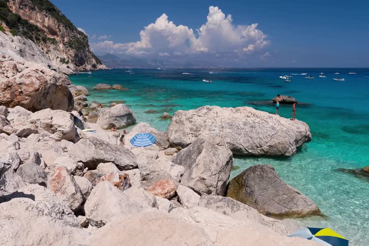 People relax and swim at cala mariolu beach in italy surrounded by large white rocks and turquoise water with boats anchored in the distance under a blue sky with white clouds