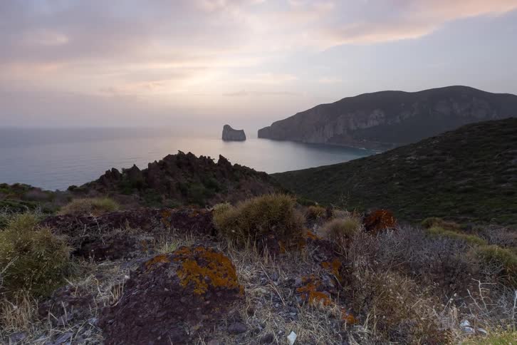 Mountains on the coast with a rock formation in the water as the sun glows in a cloudy sky